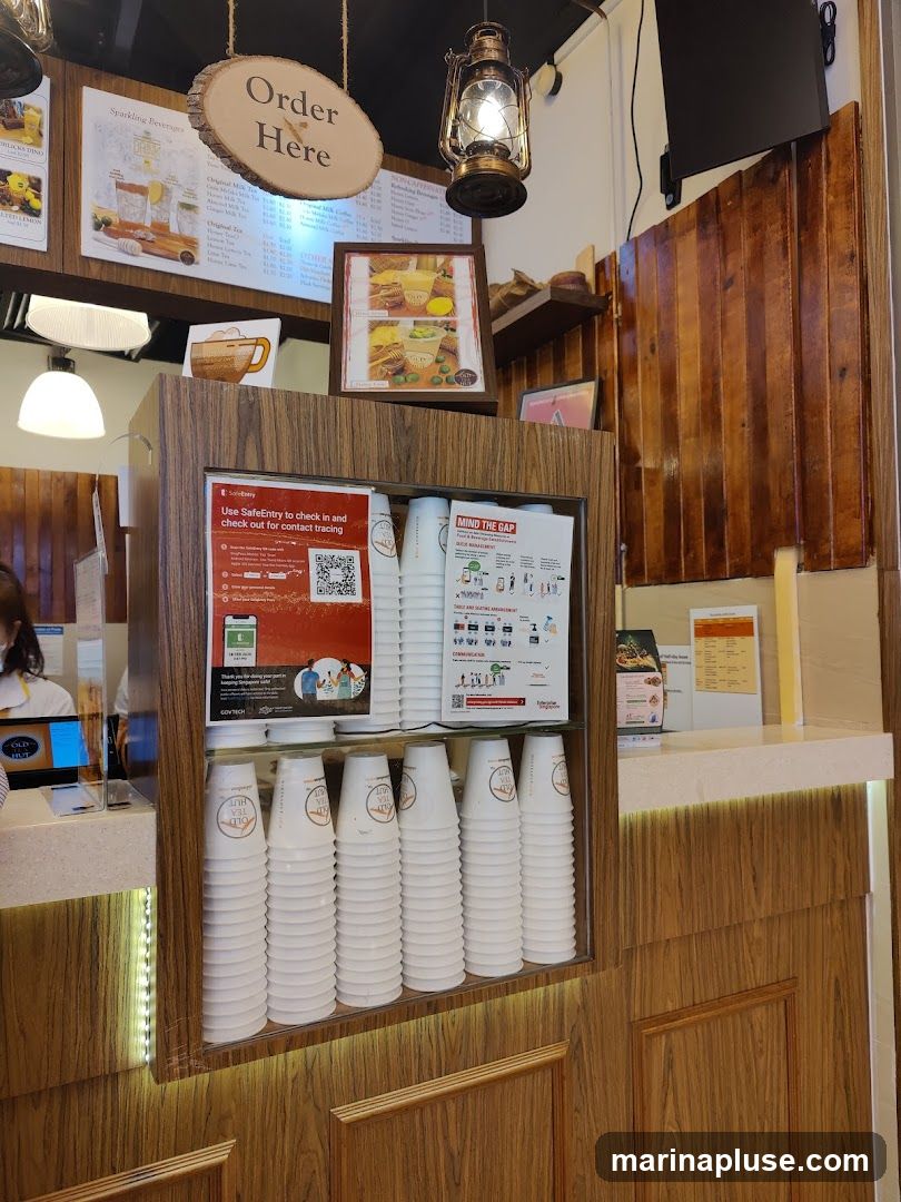 A wooden counter displays stacks of white paper cups with a logo, alongside various menus and informational posters. An "Order Here" sign hangs above.
