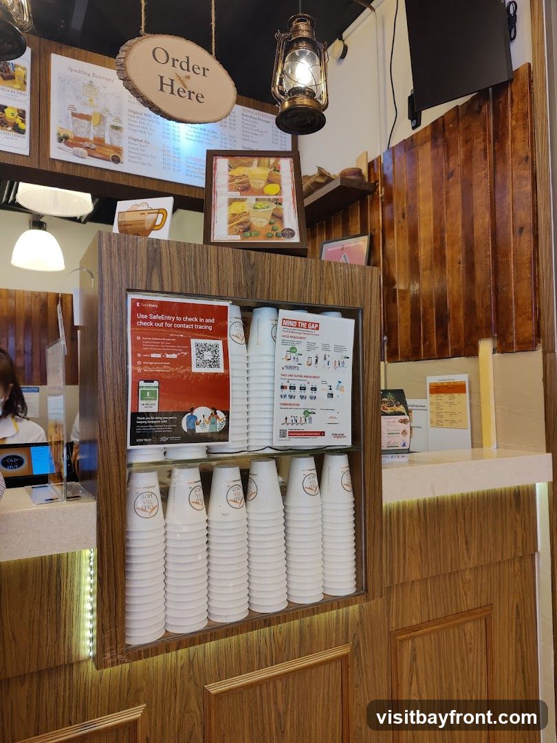 A wooden counter displays stacks of white paper cups with a logo, alongside various menus and informational posters. An "Order Here" sign hangs above.