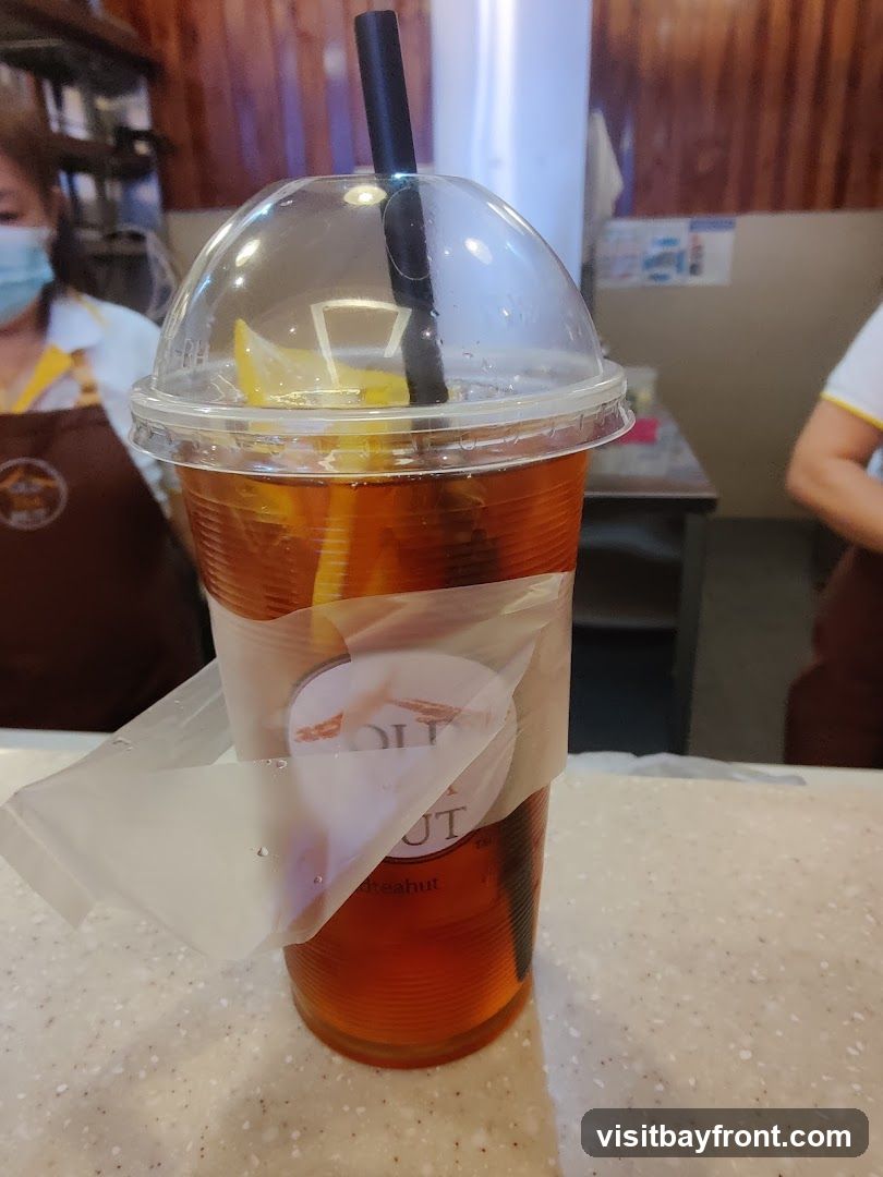 A close-up of a clear plastic cup filled with iced tea and a slice of lemon. The cup has a domed lid and a black straw. A sticker with a logo and text is partially attached to the cup. In the background, two people wearing aprons are visible, suggesting a food service environment.