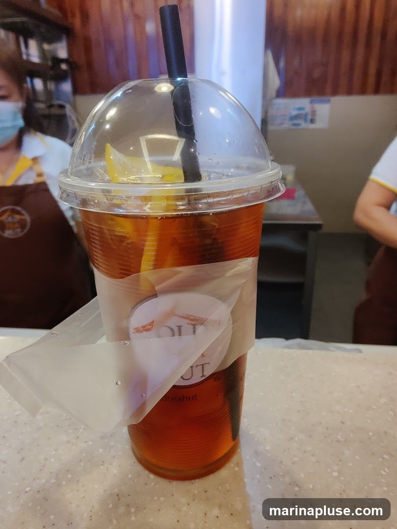A close-up of a clear plastic cup filled with iced tea and a slice of lemon. The cup has a domed lid and a black straw. A sticker with a logo and text is partially attached to the cup. In the background, two people wearing aprons are visible, suggesting a food service environment.
