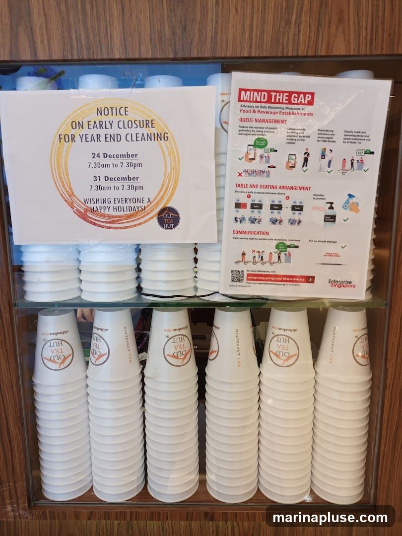 A display case filled with stacked paper cups, some with the 'Old Tea Hut' logo. Two notices are visible: one announcing early closure for year-end cleaning on December 24th and 31st, and another titled 'Mind the Gap' providing advisory on safe distancing measures for food and beverage establishments, including queue management, table and seating arrangements, and communication strategies.