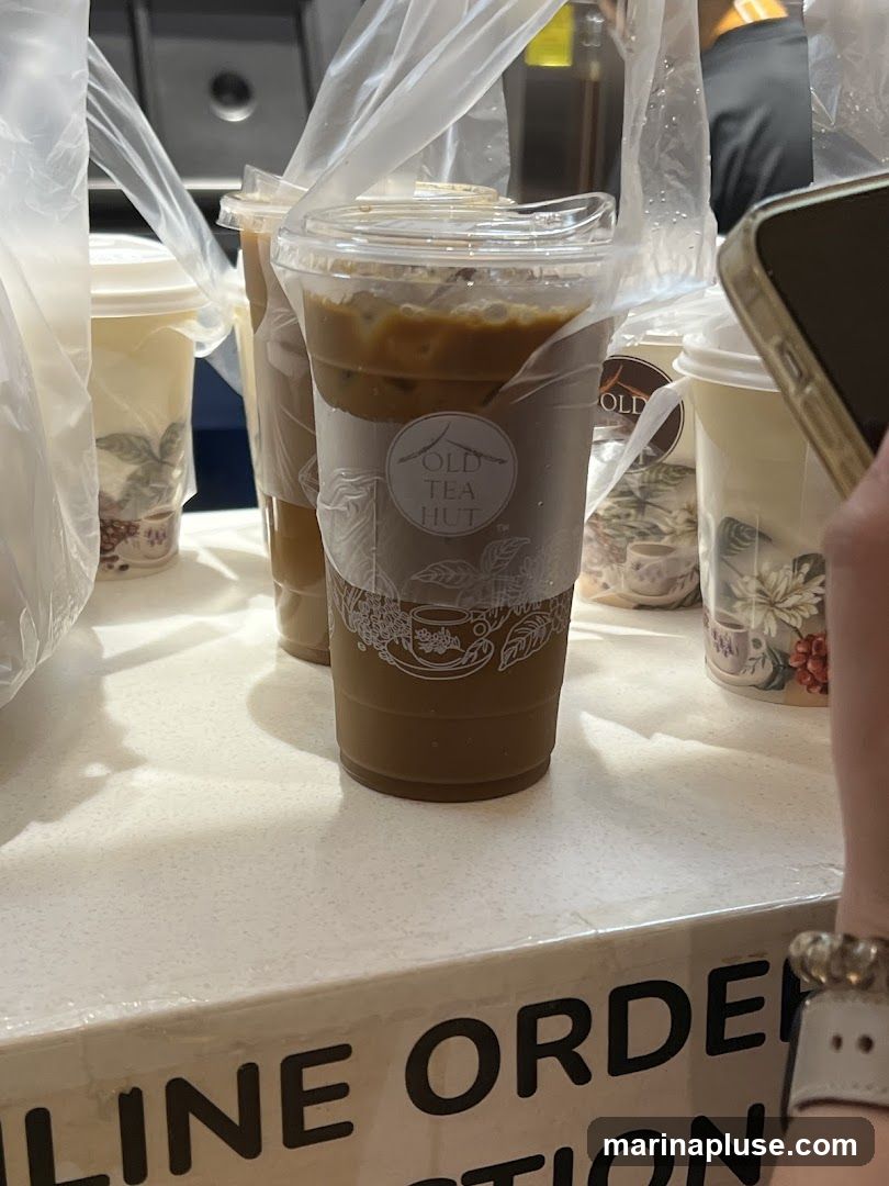 Several iced drinks in plastic cups are arranged on a counter. The cups have a logo that says "OLD TEA HUT". Some drinks are in plastic bags. A person's hand holding a phone is visible on the right side of the image. Text on the counter reads "LINE OR ORDER".