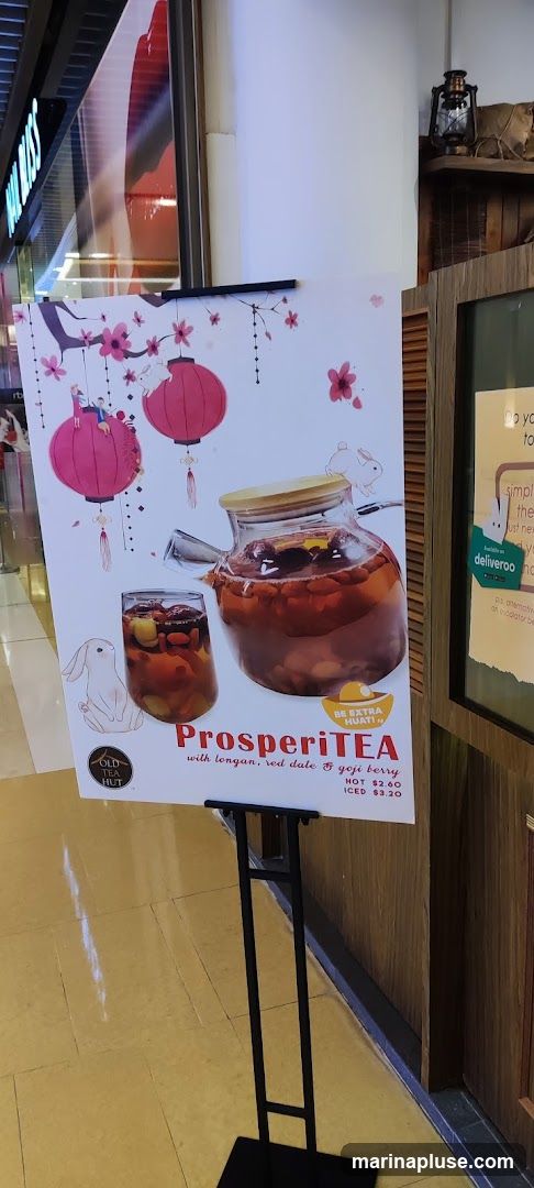A promotional poster for "ProsperiTEA" with longan, red date, and goji berry. The poster features illustrations of lanterns, cherry blossoms, and rabbits, along with images of the tea in a teapot and a glass. Prices for hot and iced versions are listed. The poster is displayed on a black easel.