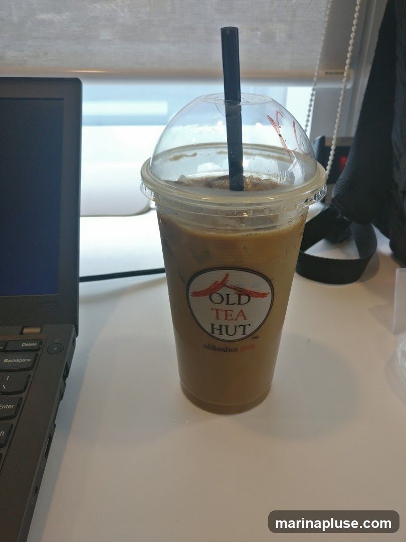 A plastic cup filled with iced coffee, topped with a domed lid and a black straw. The cup has a circular logo that reads "OLD TEA HUT" with a website address below. The cup is placed on a white surface next to a laptop.