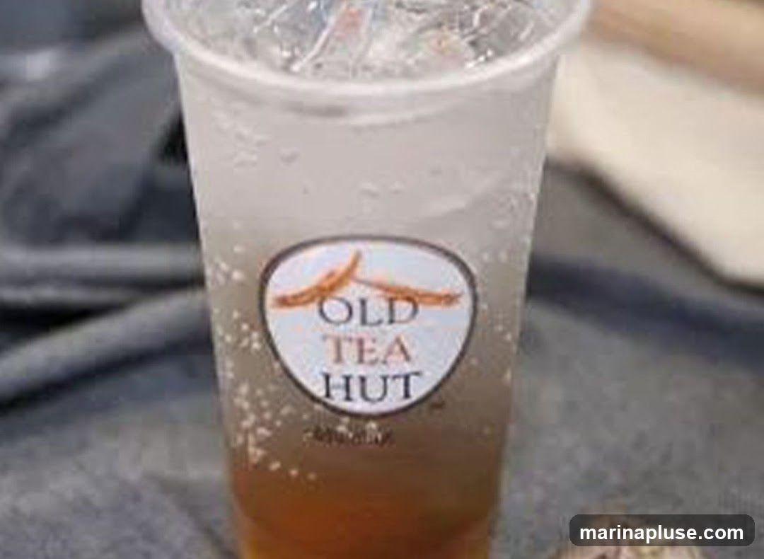 A clear plastic cup filled with a beverage, ice, and a dark liquid at the bottom. The cup has a circular logo that reads "OLD TEA HUT" with an orange design above the text. The cup is placed on a textured surface, possibly fabric.