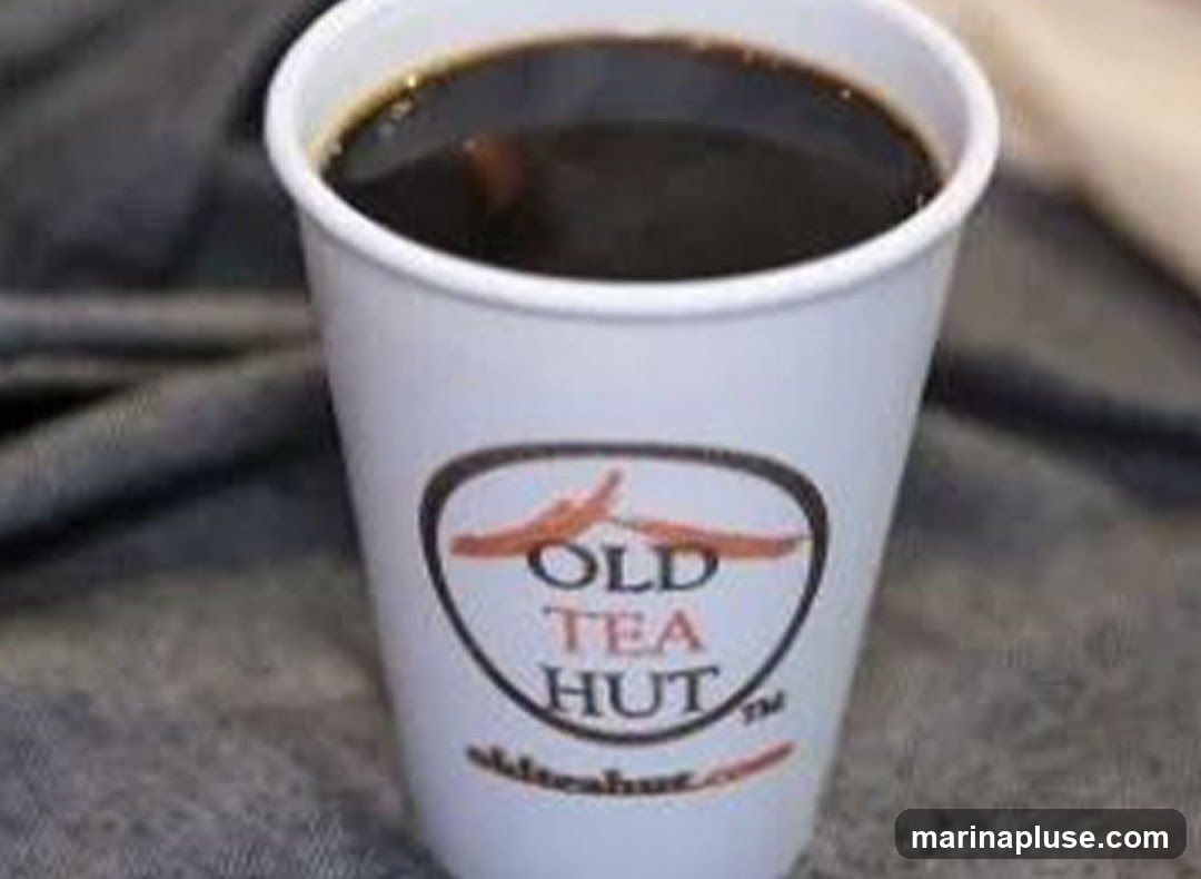 A white paper cup filled with dark coffee. The cup has a logo that reads "OLD TEA HUT" with a website address below it. The cup is placed on a textured surface.