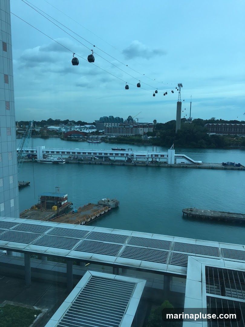 A view of a harbor with several cable cars suspended on wires overhead. In the distance, there are buildings and a tall tower. The water is calm, and there are several boats and barges visible. The foreground shows a modern walkway with a slatted roof.