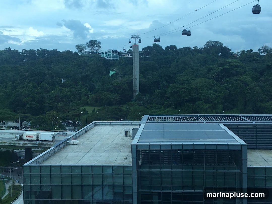 A panoramic view captures several cable cars suspended on wires, traversing over a dense, verdant forest. In the distance, buildings are visible atop the tree-covered hills. In the foreground, a modern glass-fronted building with a flat rooftop is prominent, with another rooftop extending to the left. A highway with trucks can be seen in the mid-ground, adjacent to the forested area. The sky is overcast with dramatic clouds.