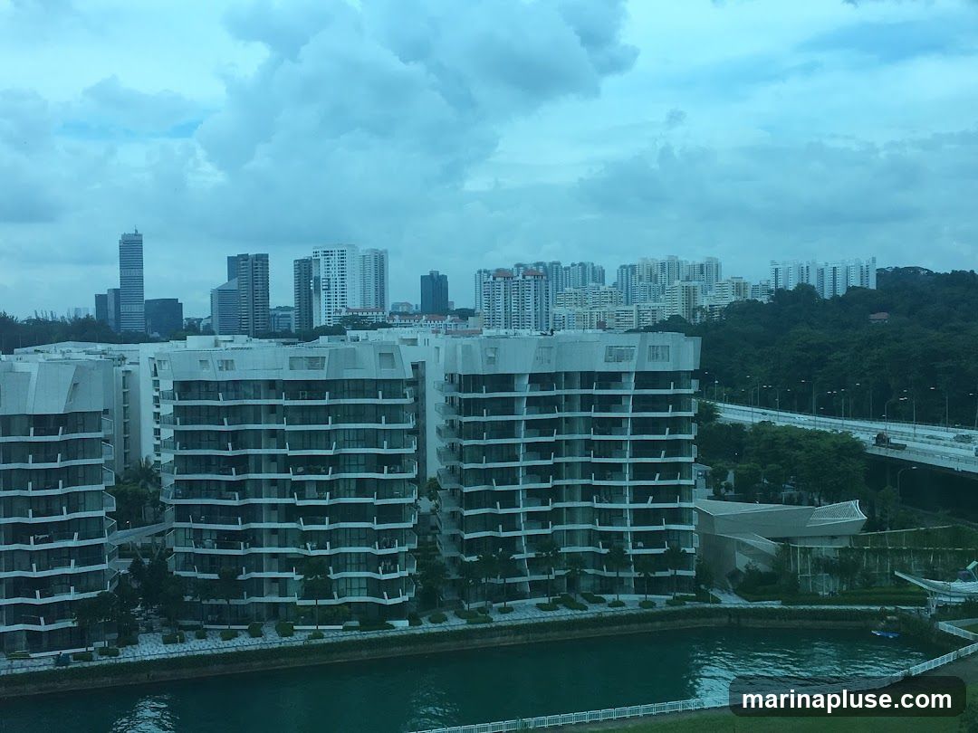 A wide shot of a cityscape featuring modern residential buildings with balconies, a waterway in the foreground, and a highway bridge in the background. The sky is overcast with dramatic clouds. The buildings are predominantly white and grey, with many windows. Lush green trees surround some of the structures and line the distant hills. The water reflects the cloudy sky and surrounding architecture.