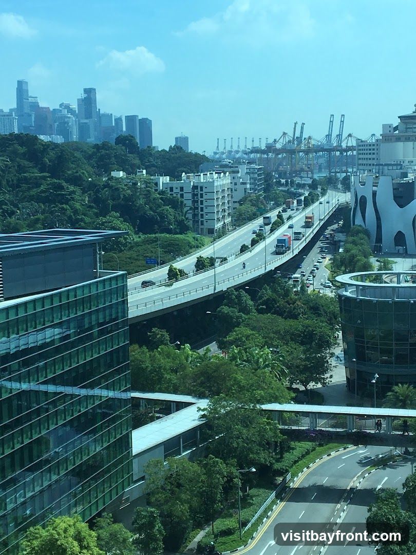 An aerial view of a modern cityscape featuring a multi-lane highway with traffic, surrounded by lush greenery and contemporary buildings. In the distance, a dense urban skyline is visible, along with numerous port cranes, suggesting a busy shipping and industrial area. The image captures a blend of urban development, infrastructure, and natural elements under a bright, partly cloudy sky.