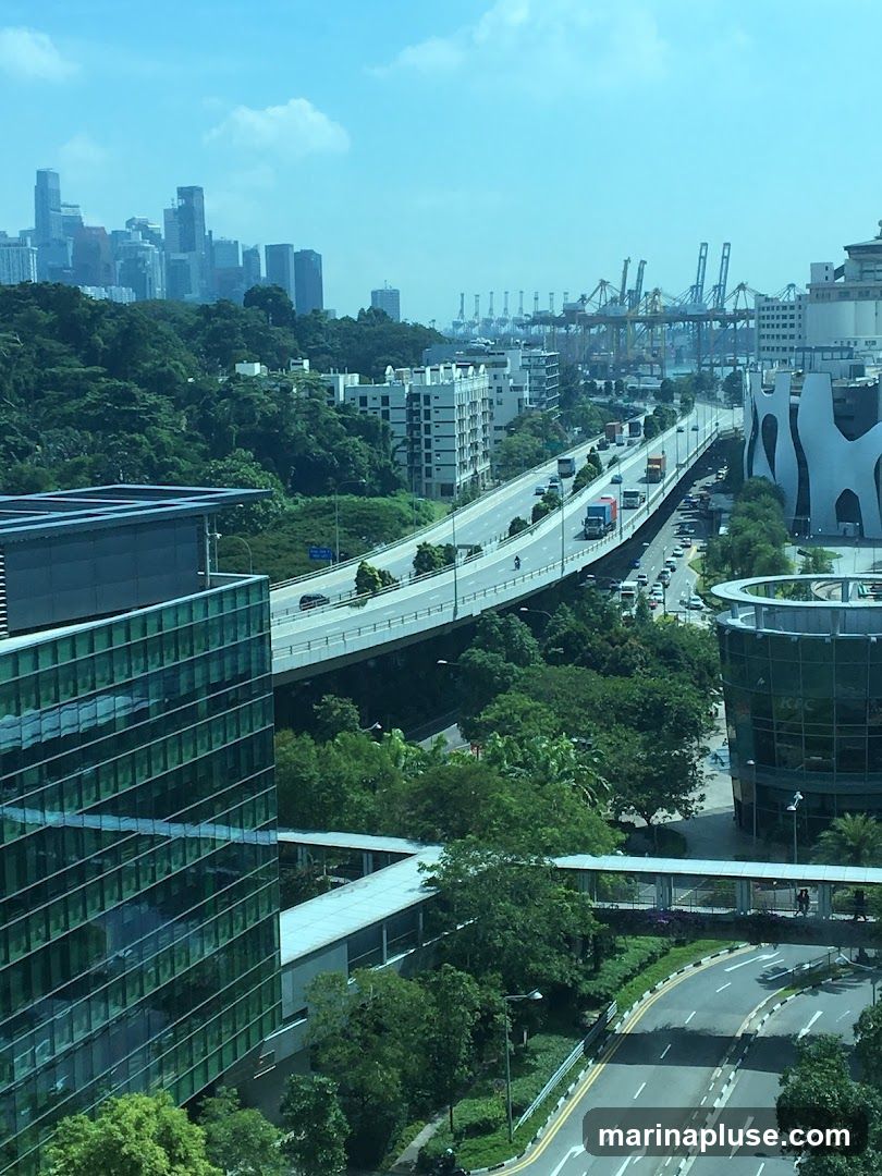 An aerial view of a modern cityscape featuring a multi-lane highway with traffic, surrounded by lush greenery and contemporary buildings. In the distance, a dense urban skyline is visible, along with numerous port cranes, suggesting a busy shipping and industrial area. The image captures a blend of urban development, infrastructure, and natural elements under a bright, partly cloudy sky.