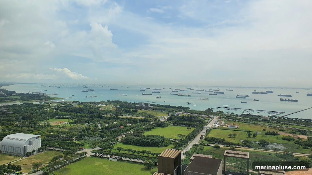 A wide, high-angle shot captures a vast expanse of ocean dotted with numerous cargo ships and smaller vessels. The water is a calm blue-green, meeting a hazy horizon under a partly cloudy sky. In the foreground, a lush green landscape unfolds, featuring manicured lawns, scattered trees, and several modern buildings. A prominent, large, grey, arched-roof building is visible on the left, while a complex of darker buildings sits closer to the coastline. A long, man-made pier or breakwater extends into the water on the right side of the frame. The scene suggests a bustling port or a significant maritime traffic area with adjacent urban development and green spaces.