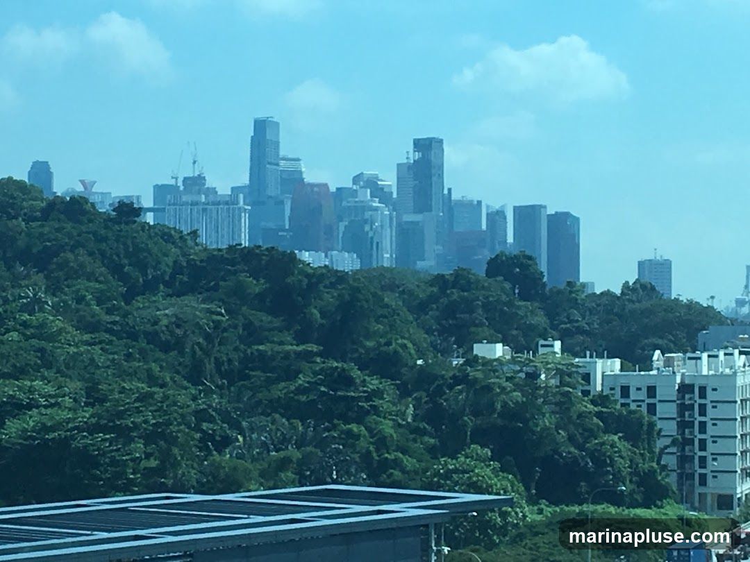 A panoramic view of a city skyline dominated by modern skyscrapers, set against a backdrop of lush green hills and trees. The sky is a clear blue with scattered white clouds. In the foreground, a portion of a modern building with a dark, slatted roof is visible. The overall scene suggests an urban environment integrated with natural landscapes.