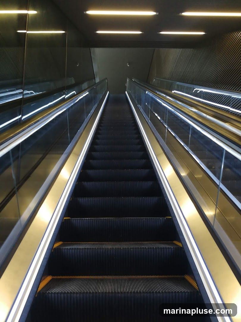 A view looking up a modern escalator with glass railings and bright lighting. The escalator steps are dark and ribbed, with yellow accents at the edges. The surrounding walls are dark, with reflective surfaces and ribbed textures. Overhead, fluorescent lights illuminate the space.