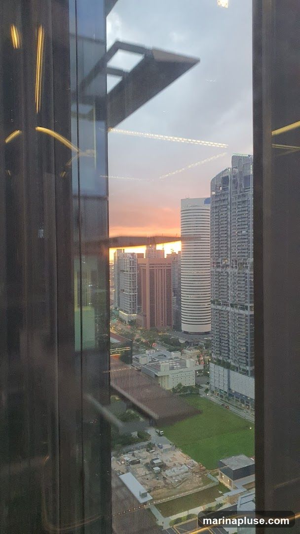 A view of a city skyline at dusk, seen through a window. The sky is painted with warm hues of orange and pink from the setting sun. Several tall buildings of varying architectural styles dominate the landscape, with some appearing modern and sleek, while others have a more traditional design. Below, a large green field is visible, with some construction sites and roads interspersed. Reflections of interior lights and architectural elements are visible on the window glass, adding depth to the scene.