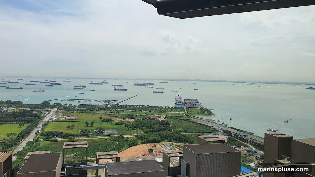 An aerial view captures a vast expanse of water dotted with numerous cargo ships and a large cruise ship docked at a pier. In the foreground, a coastal landscape features green fields, roads, and construction sites. The sky is overcast with scattered clouds.