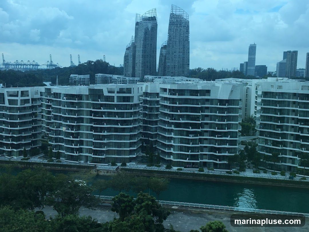 A panoramic view of several modern apartment buildings with white facades and numerous balconies, situated along a tranquil blue waterway. In the background, a cityscape with taller skyscrapers and industrial cranes can be seen under a cloudy sky. The scene suggests an urban waterfront development.