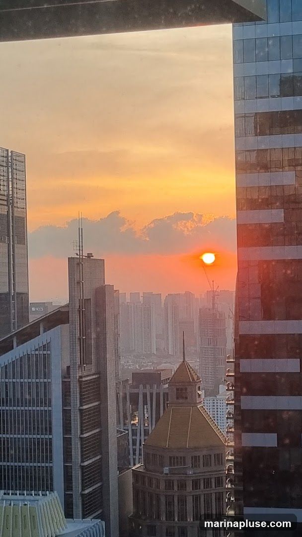 A vibrant sunset paints the sky with hues of orange and yellow, casting a warm glow over a dense cityscape. Tall modern buildings with glass facades dominate the foreground, reflecting the sky's colors. In the distance, a hazy layer of buildings stretches towards the horizon, with a few construction cranes visible. The sun, a bright orange orb, is partially obscured by wispy clouds.
