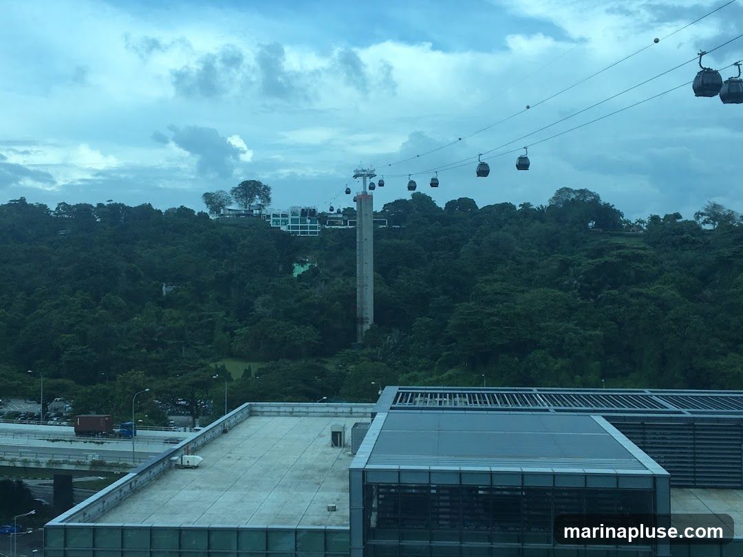 A view of cable cars suspended on wires, traversing over a dense, green forested hill. In the foreground, a modern building with a flat rooftop is visible, alongside a highway with a truck. The sky is overcast with dramatic clouds.