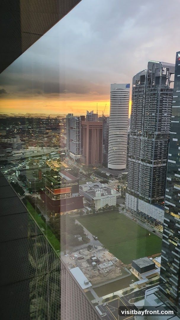 An aerial view of a cityscape at sunset, with a vibrant orange and yellow sky visible above a dense collection of modern skyscrapers and buildings. The foreground shows a large green field, with some construction sites and roads visible. The reflection of a patterned surface is visible on the left side of the image.