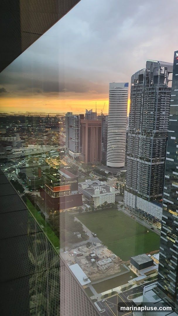 An aerial view of a cityscape at sunset, with a vibrant orange and yellow sky visible above a dense collection of modern skyscrapers and buildings. The foreground shows a large green field, with some construction sites and roads visible. The reflection of a patterned surface is visible on the left side of the image.
