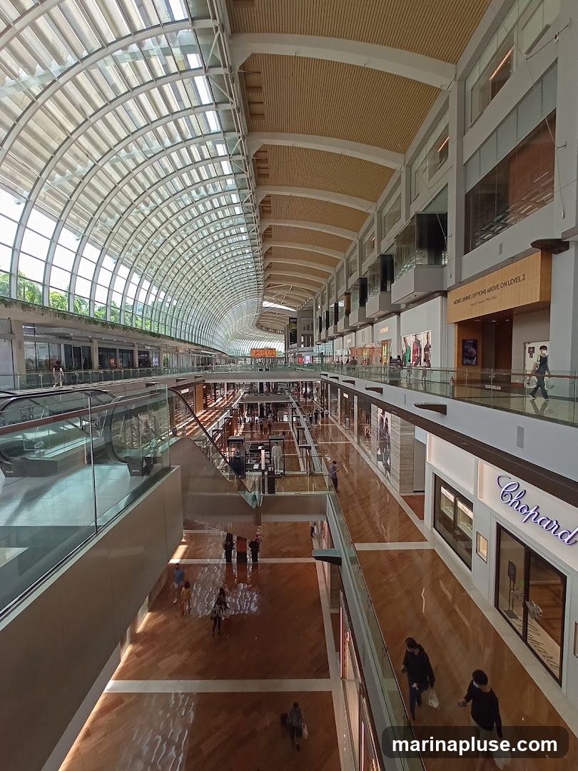 A wide-angle view of the interior of a large, modern shopping mall. The architecture features a curved glass roof, wooden floors, and multiple levels of shops. People are seen walking around the mall, and escalators are visible. A Chopard store is identifiable on the right side.