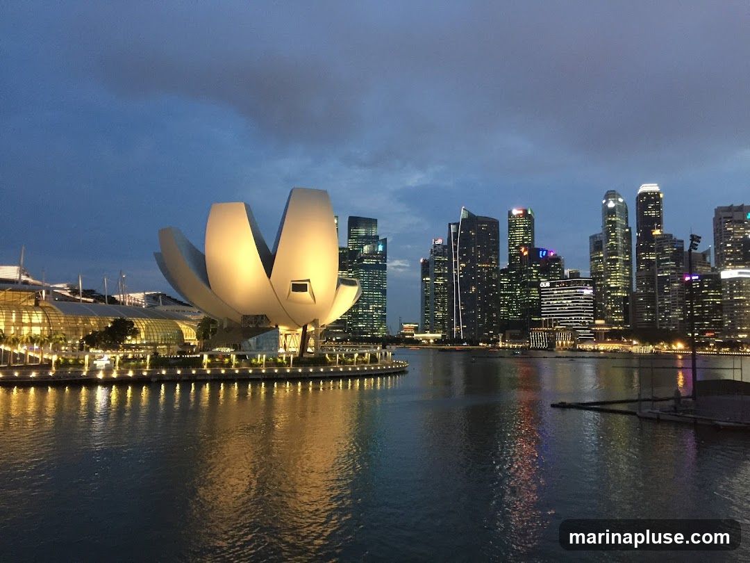 A wide shot of the Singapore skyline at dusk, with the ArtScience Museum in the foreground and numerous skyscrapers in the background. The water in front reflects the lights of the city.