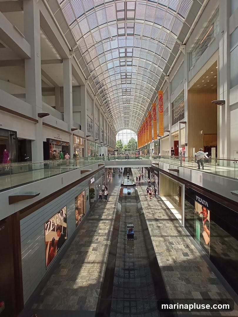 A long, wide shopping mall interior with a high, arched glass ceiling. Sunlight streams through the ceiling, casting long shadows on the tiled floor. Shops line both sides of the mall, with some displaying large advertisements. People are seen walking and browsing.