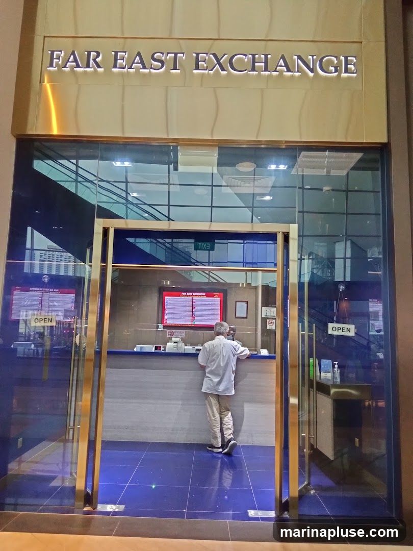 The exterior of a currency exchange shop with a large sign above the entrance that reads "FAR EAST EXCHANGE". The shop has glass doors and windows, with a person standing at the counter inside. There are also signs indicating that the shop is open.