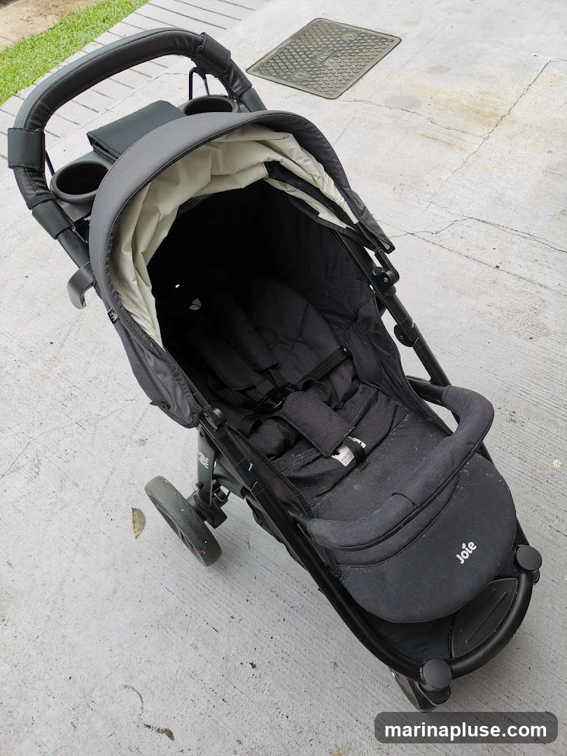A dark gray Joie stroller is shown from a high angle, resting on a concrete surface. The stroller has a canopy, seat with harness, and a handle with cup holders. The brand name "Joie" is visible on the seat cushion.