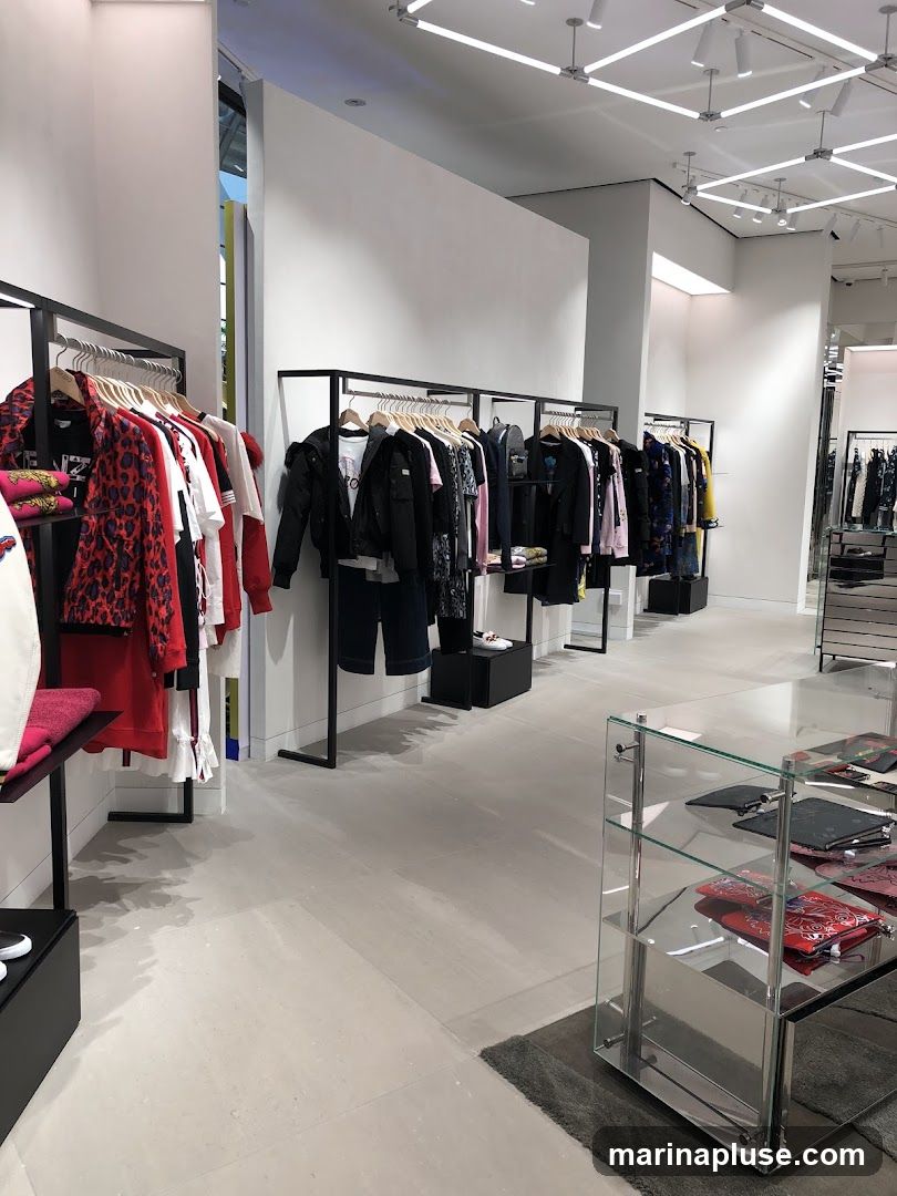 A well-lit clothing store interior with racks of apparel. On the left, racks display women's clothing with bold patterns and bright colors, including red and leopard print. In the center and right, racks showcase a collection of jackets, shirts, and pants, predominantly in darker tones. A glass display case on the right exhibits various accessories, such as clutches and wallets. The store features a modern design with minimalist fixtures and a neutral color palette for the walls and flooring.