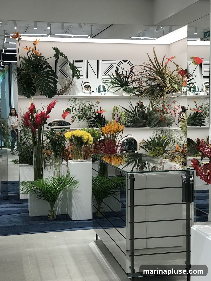 The image shows the interior of a Kenzo store, featuring a prominent display of various plants and flowers arranged on shelves and in vases. The Kenzo Paris logo is visible on the back wall. Several mannequins or display heads wearing helmets are also present on the shelves. A mirrored display case is in the foreground, reflecting the store's interior. A woman is visible on the left side of the image, near the entrance.