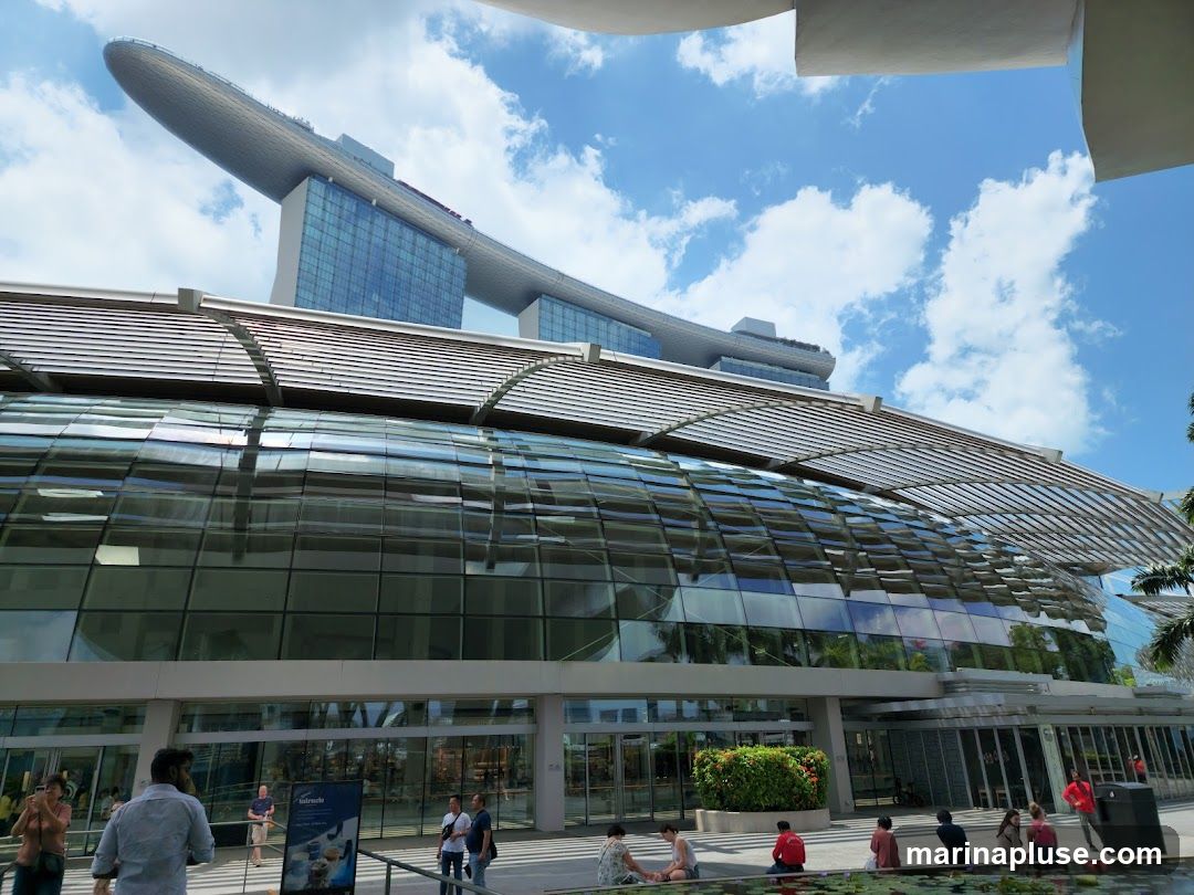 A wide shot of the Marina Bay Sands complex in Singapore on a sunny day with a blue sky and scattered clouds. The iconic boat-shaped structure is visible in the background, perched atop three tall buildings. In the foreground, a modern building with a curved glass facade reflects the sky and surrounding greenery. People are seen walking and sitting outside the building. A sign with text and images is placed near the entrance.