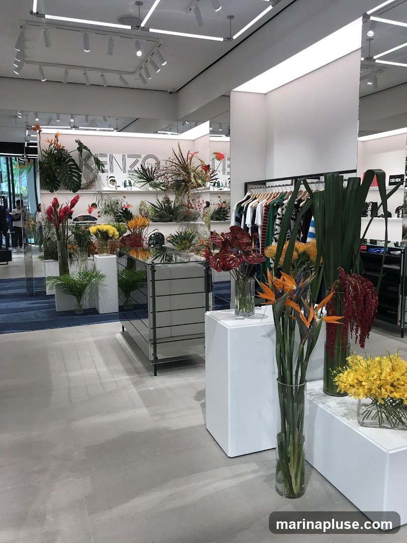 The image shows the interior of a modern store. In the foreground, there are several large floral arrangements in glass vases, including Bird of Paradise flowers and yellow orchids. White pedestals are used to display the flowers. In the mid-ground, there are clothing racks with various garments, and a display case with mirrored surfaces. In the background, a wall features a large decorative element with the word "KENZO" in a stylized font, surrounded by more floral displays and merchandise. The store has a clean, minimalist aesthetic with white walls and ceiling, and track lighting.