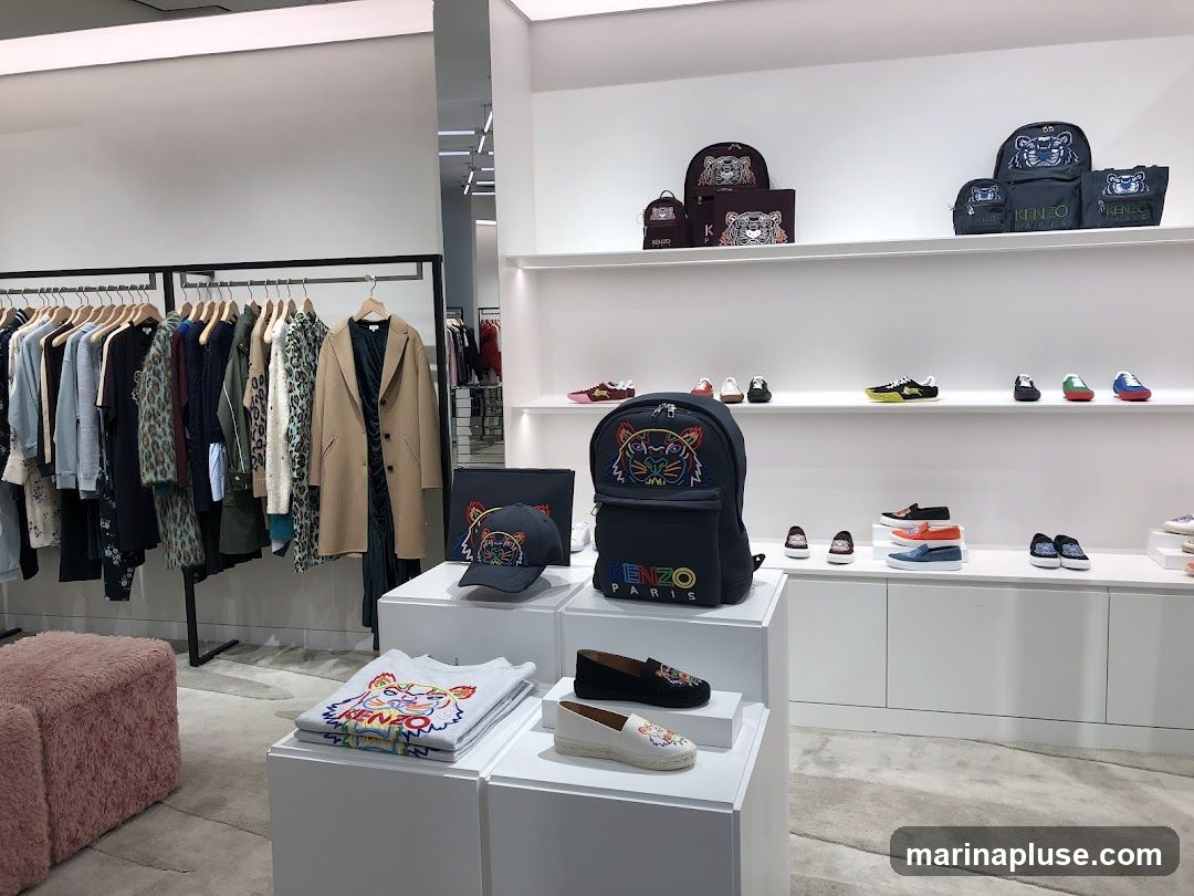 A retail store display featuring Kenzo branded clothing and accessories. On the left, racks of clothing are displayed, including coats, sweaters, and patterned garments. In the center and right, shelves and display stands showcase backpacks, handbags, hats, t-shirts, and shoes, many featuring the iconic Kenzo tiger motif and logo. The store has a clean, modern aesthetic with white shelving and displays.