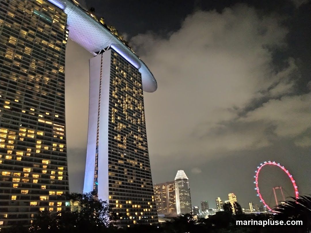 A nighttime view of the Marina Bay Sands hotel with its iconic Skypark, illuminated by numerous lights. To the right, the Singapore Flyer Ferris wheel is visible, also lit up. The cityscape of Singapore is in the background under a cloudy sky.