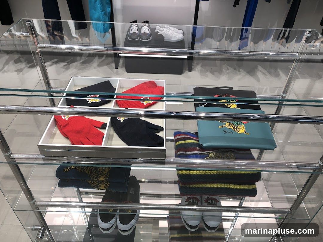 A glass display shelf showcases various accessories and footwear. On the top shelf, there are two pairs of sneakers. The second shelf displays hats and gloves in red and black, each with a logo. The third shelf holds a black clutch and a light blue pouch with a tiger design and the word "Paris" printed on it, along with a striped scarf. The bottom shelf displays more striped scarves and two pairs of sneakers.