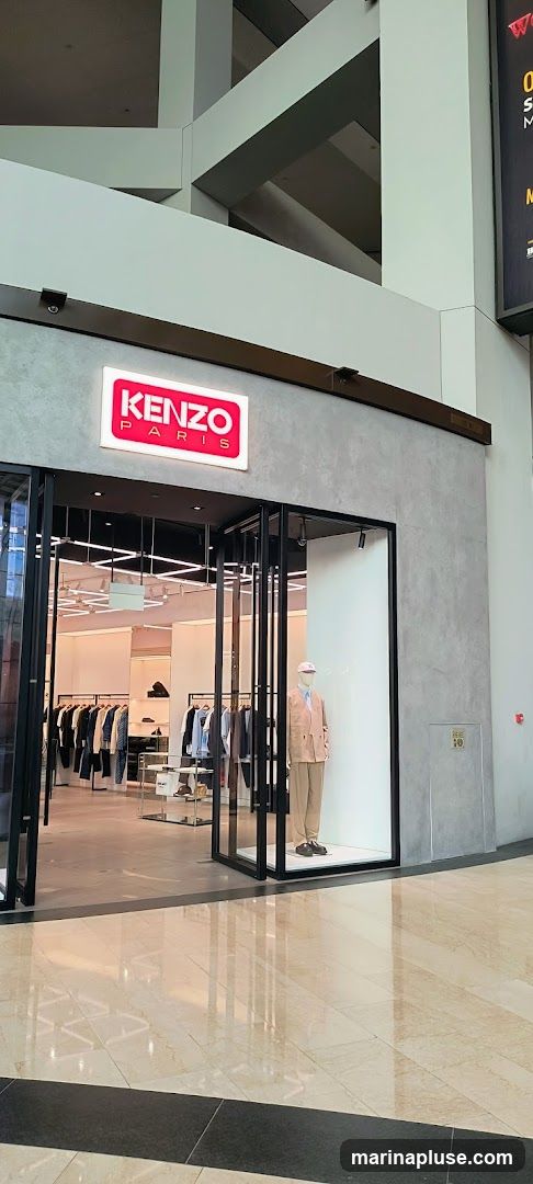 The exterior of a Kenzo Paris store. A prominent illuminated sign with the KENZO PARIS logo is visible above the entrance. The store features a large glass display window showcasing a mannequin dressed in a light-colored suit. Inside, racks of clothing are displayed.