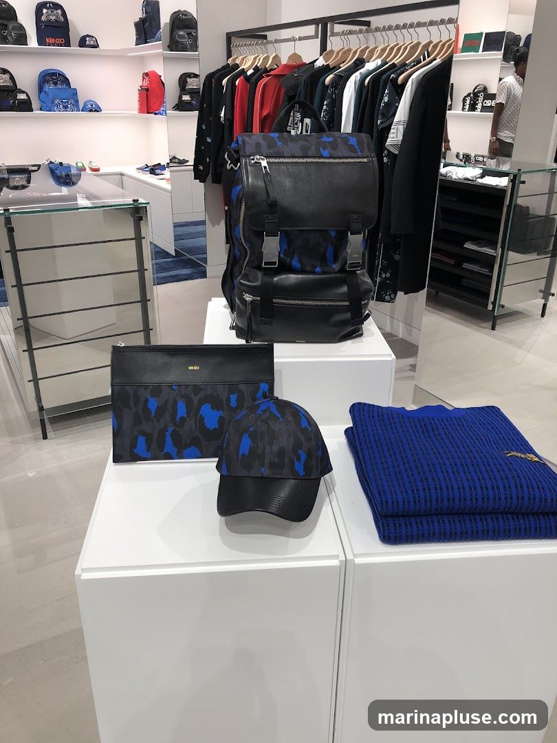A retail display featuring a black backpack with a blue and black camouflage pattern, a matching clutch bag, a baseball cap with a similar pattern and black leather brim, and a folded blue waffle-knit sweater. In the background, clothing racks with various garments and shelves stocked with bags and accessories are visible.