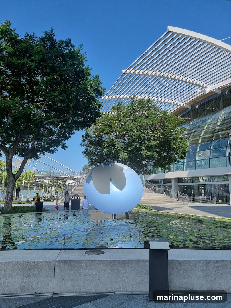 A large, modern building with a distinctive slatted roof and glass facade is visible under a clear blue sky. In front of the building, a serene water feature with lily pads and purple flowers reflects the surroundings. A large, highly reflective sphere sculpture is positioned in the water, mirroring the sky and the architecture. Several people are seen in the area, some sitting near the water and others walking towards the building's entrance. The scene conveys a sense of contemporary design and public space.