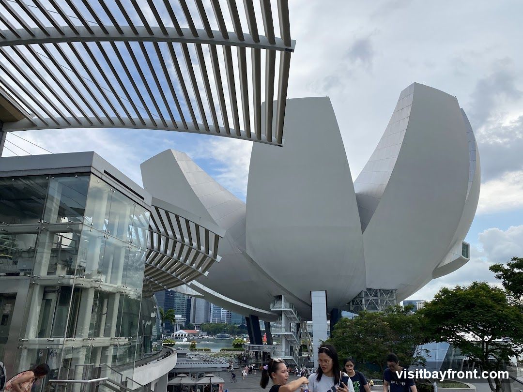 A wide shot of a modern building with a distinctive petal-like structure, set against a cloudy sky. In the foreground, a group of people are walking down stairs. The building features glass elements and a metallic exterior.