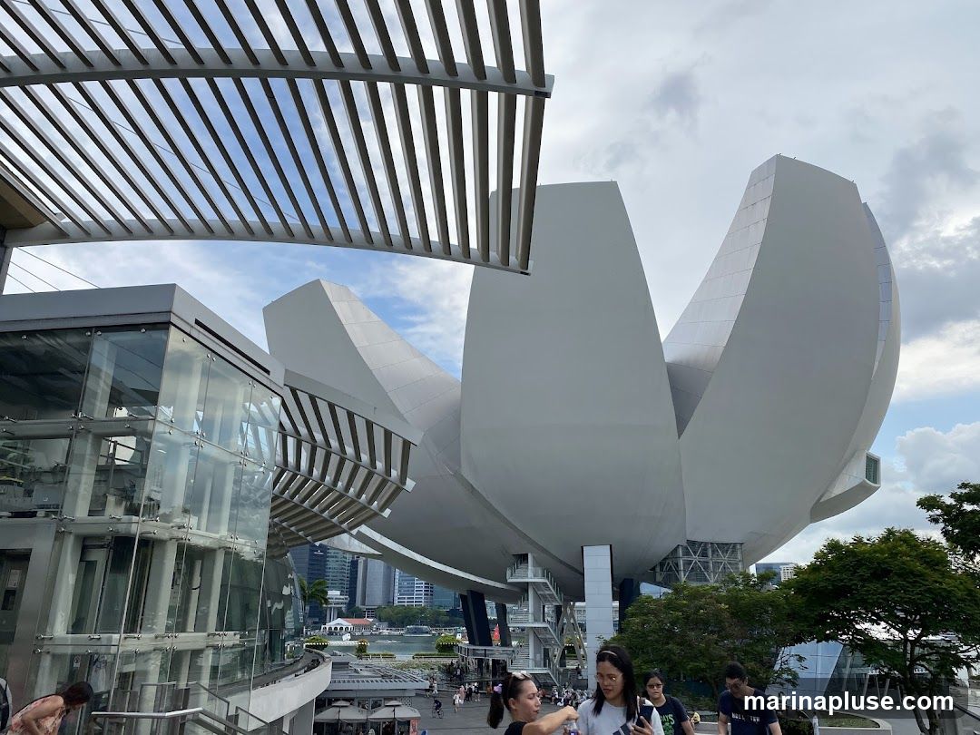 A wide shot of a modern building with a distinctive petal-like structure, set against a cloudy sky. In the foreground, a group of people are walking down stairs. The building features glass elements and a metallic exterior.