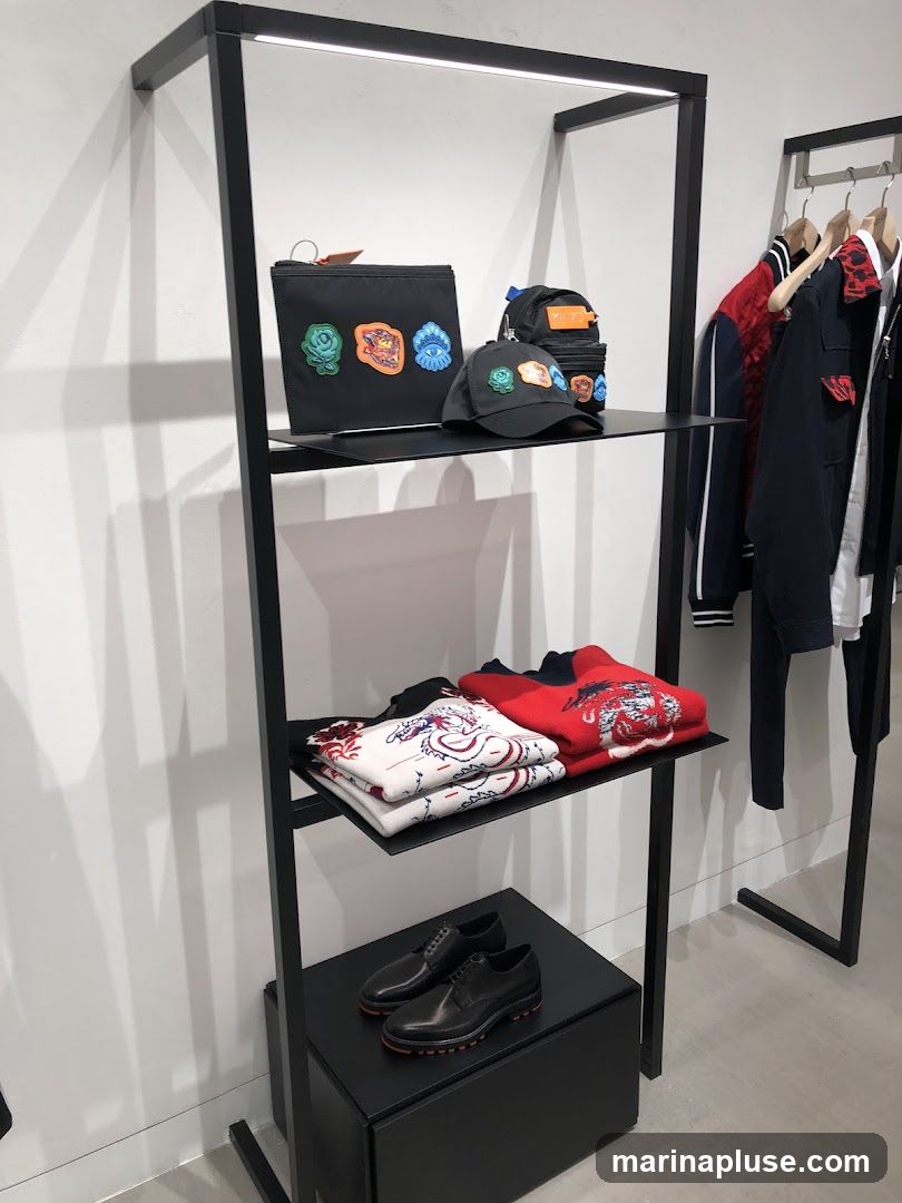 A retail display shelf with various clothing items and accessories. On the top shelf, there is a black pouch with patches, a black baseball cap with patches, and a small backpack. The middle shelf holds folded sweaters, one white with red and blue dragon designs, and one red with similar dragon designs. On the bottom shelf, a pair of black leather dress shoes with red soles is displayed. To the right, clothing items are hanging on a rack, including jackets and shirts.