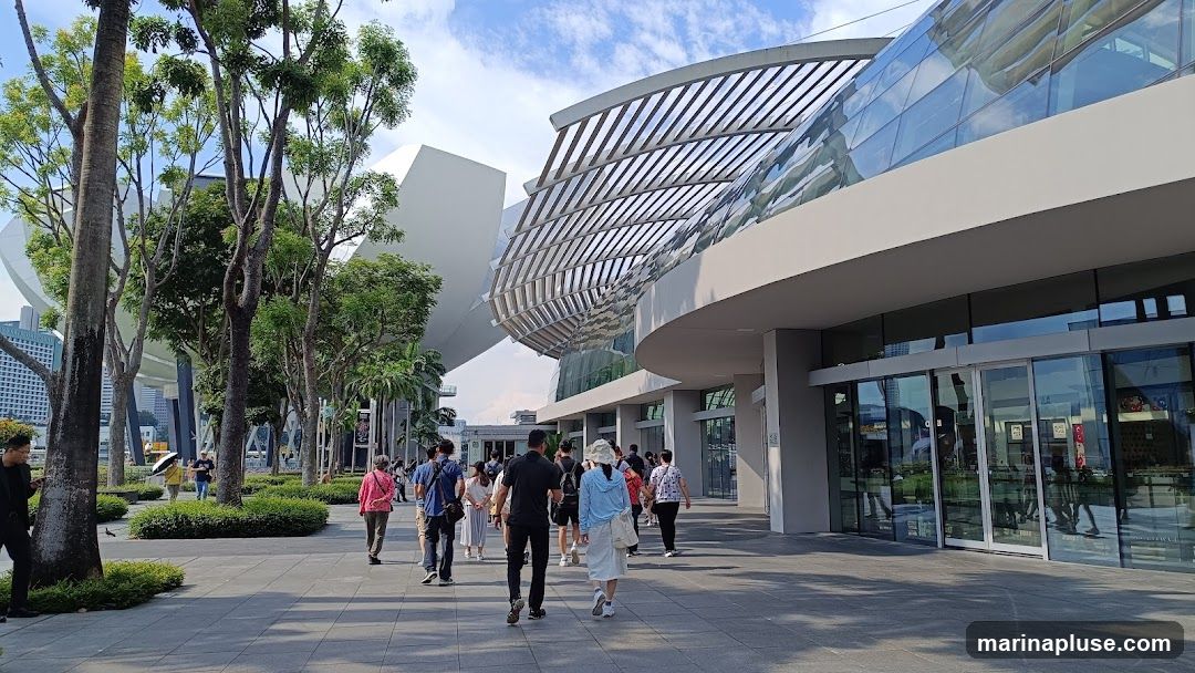 A wide shot of a modern building with a curved glass facade and a slatted roof structure. People are walking along a paved walkway in front of the building, which is lined with trees and greenery. The sky is blue with scattered clouds.