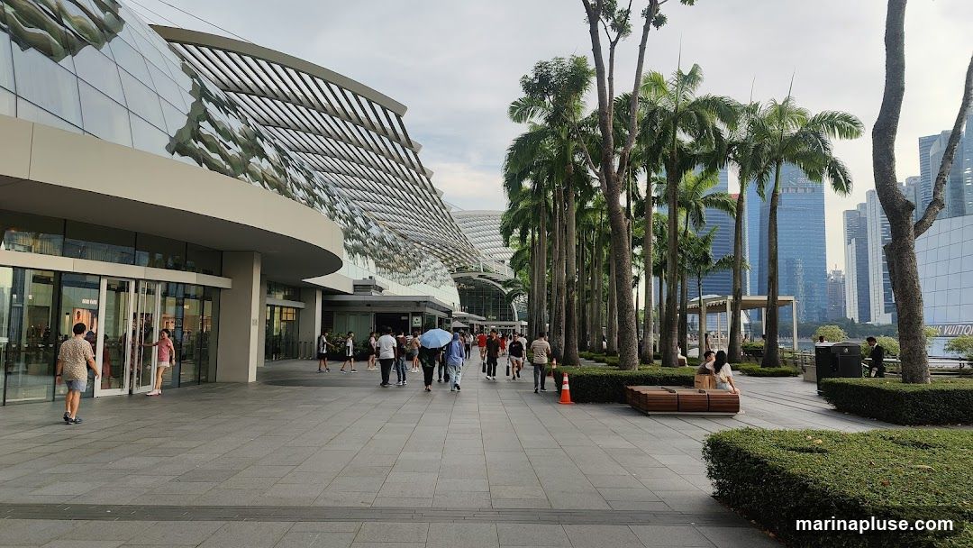 A wide shot of a modern shopping mall with a distinctive curved glass facade and a slatted roof. A paved walkway in front of the mall is lined with tall palm trees and people strolling. In the background, several skyscrapers are visible, suggesting an urban setting. A sign that appears to say "LOUIS VUITTON" is visible on a building in the far right background.