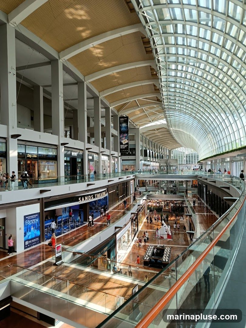 The image shows the interior of a multi-level shopping mall with a distinctive arched glass ceiling. Sunlight streams through the ceiling, illuminating the polished wooden floors and glass railings of the walkways. Shops are visible on multiple levels, with some storefronts displaying advertisements. People are seen browsing and walking along the different levels. A prominent sign reads "SANDS THEATRE". There is also a poster for "FROZEN" and a display that appears to be for a luxury brand.