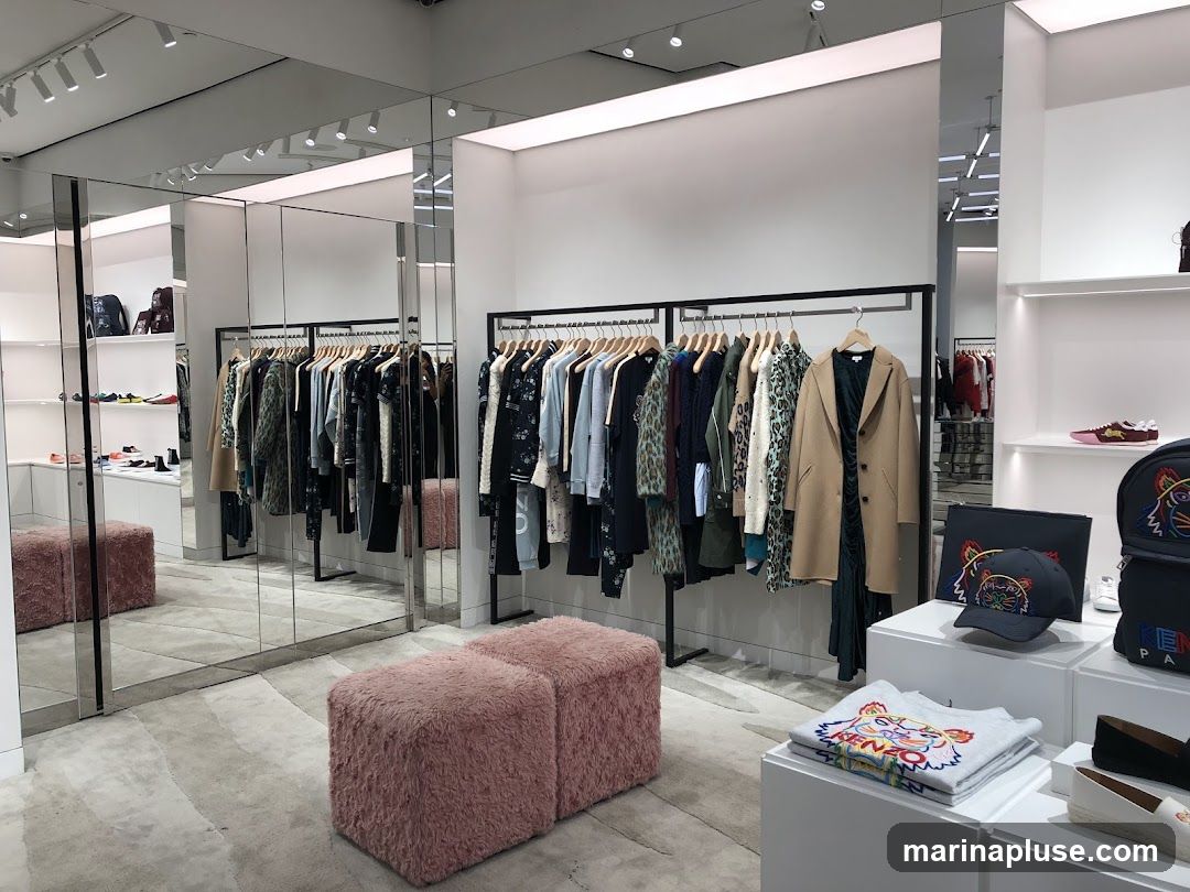 A well-lit clothing store interior with racks of apparel, shelves displaying accessories, and plush seating. The merchandise includes coats, sweaters, t-shirts, hats, bags, and shoes. Mirrors line one wall, reflecting the displays and creating a sense of spaciousness.