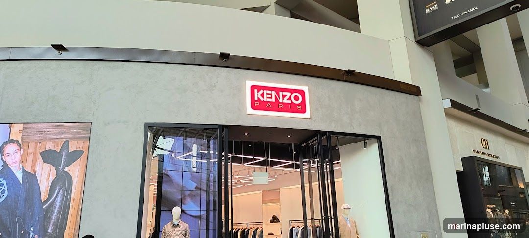 The storefront of a Kenzo Paris store is visible, featuring a prominent illuminated sign with the brand name. The entrance has large glass doors and windows, revealing the interior display of clothing on mannequins and racks. To the left of the Kenzo store, a large advertisement or display features a person in a dark outfit. To the right, another store with a sign reading 'CH CAROLINA HERRERA' is partially visible.