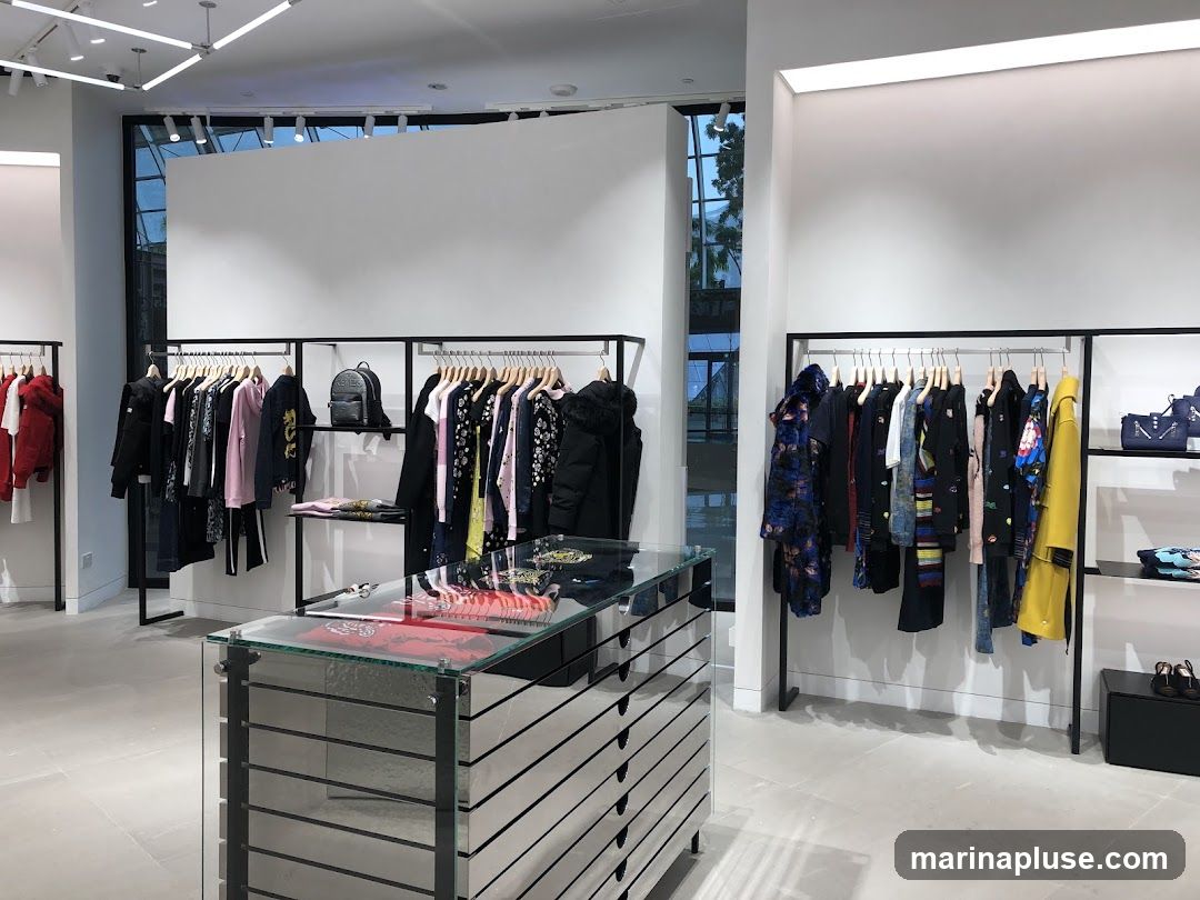A well-lit clothing store interior with racks of women's apparel. Garments include dresses, jackets, and tops in various colors and patterns. A mirrored display case with glass top is in the foreground. Shelving units display handbags and shoes.