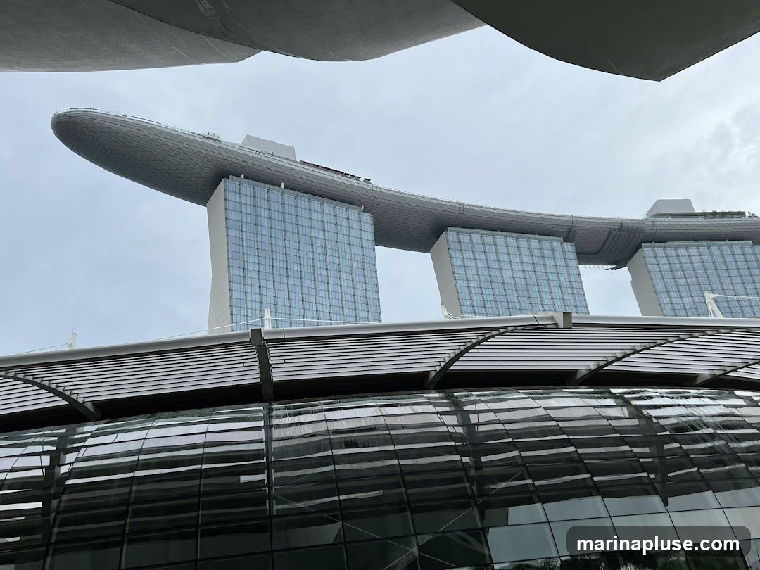 A low-angle shot of the Marina Bay Sands hotel in Singapore, with its distinctive boat-shaped rooftop and three towers. The foreground shows a modern architectural structure with a glass facade and a slatted roof. The sky is overcast.